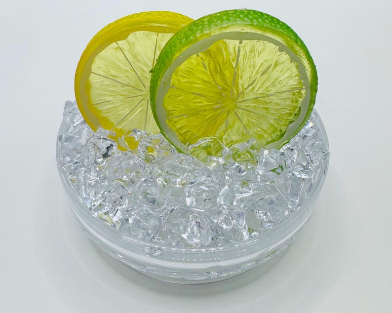 Pu&ograve; includere: Una ciotola di vetro trasparente riempita con piccoli cubetti di ghiaccio in acrilico trasparenti e irregolari. Due fette di frutta artificiale, un limone giallo brillante e un lime verde vivace, sono posizionate verticalmente, parzialmente immerse.
