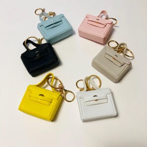 Könnte beinhalten: Eine Sammlung von Miniatur-Handtaschen-Schlüsselanhängern in verschiedenen Farben, darunter Hellblau, Rosa, Schwarz, Beige, Gelb und Weiß. Jeder Schlüsselanhänger hat einen goldfarbenen Ring und Verschluss sowie einen kleinen Griff.