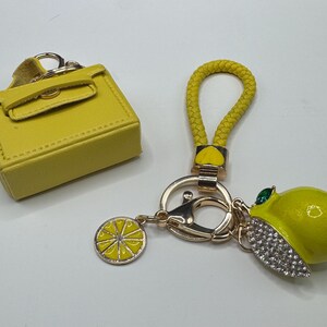 Pu&ograve; includere: Un accessorio per borsa in miniatura giallo e un portachiavi color oro con cinturino intrecciato giallo, un ciondolo a forma di fetta di limone e un limone tridimensionale decorato con cristalli trasparenti e una pietra verde.