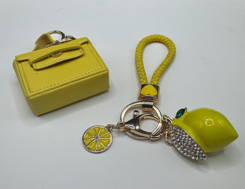 Pu&ograve; includere: Un accessorio per borsa in miniatura giallo e un portachiavi color oro con cinturino intrecciato giallo, un ciondolo a forma di fetta di limone e un limone tridimensionale decorato con cristalli trasparenti e una pietra verde.