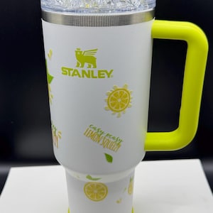 Pu&ograve; includere: Un bicchiere Stanley bianco con manico e base giallo brillante, decorato con fette di limone gialle e foglie verdi. Il testo 'easy peasy LEMON SQUEEZY' &egrave; stampato sul lato. Il coperchio presenta finti cubetti di ghiaccio, una fetta di lime e una cannuccia.