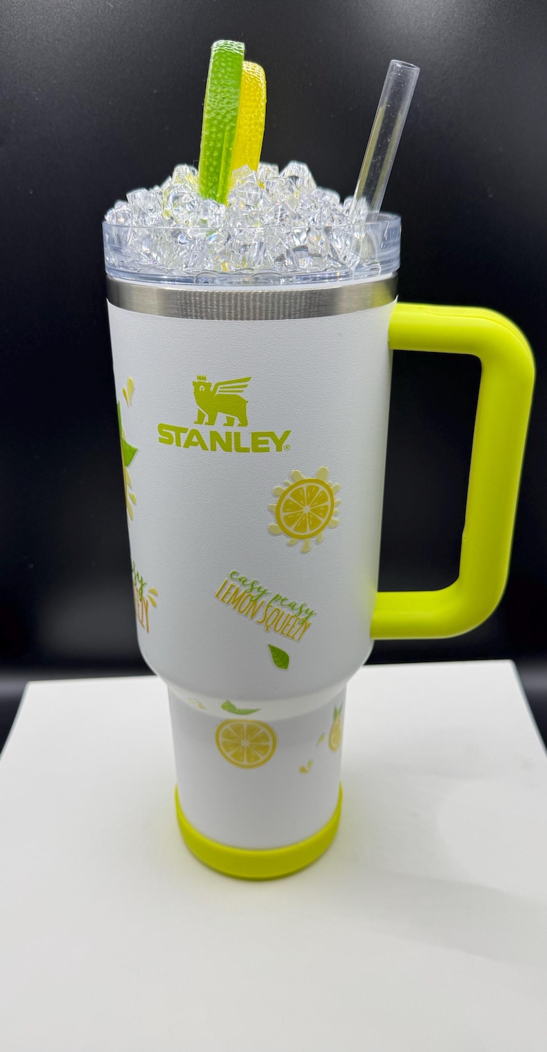 Pu&ograve; includere: Un bicchiere Stanley bianco con manico e base giallo brillante, decorato con fette di limone gialle e foglie verdi. Il testo 'easy peasy LEMON SQUEEZY' &egrave; stampato sul lato. Il coperchio presenta finti cubetti di ghiaccio, una fetta di lime e una cannuccia.