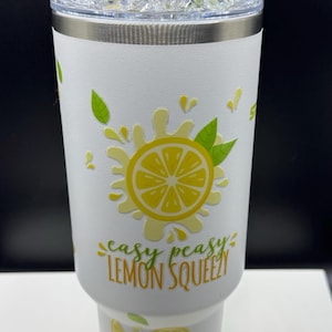 Pu&ograve; includere: Un bicchiere bianco con una grafica di limone giallo con foglie verdi e il testo "easy peasy LEMON SQUEEZY". Il coperchio &egrave; sormontato da finti cubetti di ghiaccio trasparenti e due fette di agrumi, una gialla e una verde. La base ha un rivestimento in silicone verde lime.