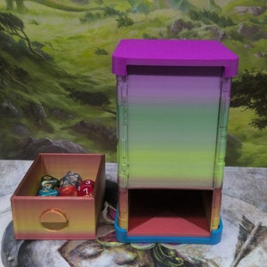 May include: A multi-colored dice tower and matching dice box. The tower has a gradient of colors, transitioning from purple to green. The dice box is rectangular with a circular opening. Several dice are visible inside the box.