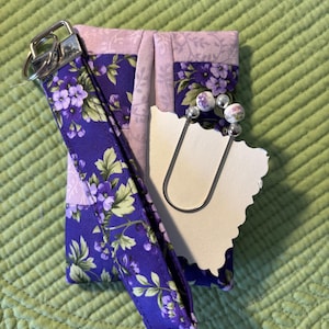 May include: A fabric wristlet keychain featuring a purple floral pattern with green leaves, paired with a silver metal clasp. Attached is a large silver safety pin adorned with two floral beads and a blank, scalloped-edge white paper tag.