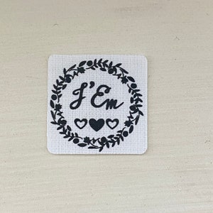 Puede incluir: Un parche cuadrado de tela blanca con texto negro y una corona decorativa. El texto dice "J'Em" dentro de una corona de hojas, con tres corazones debajo. El parche tiene una superficie texturizada.
