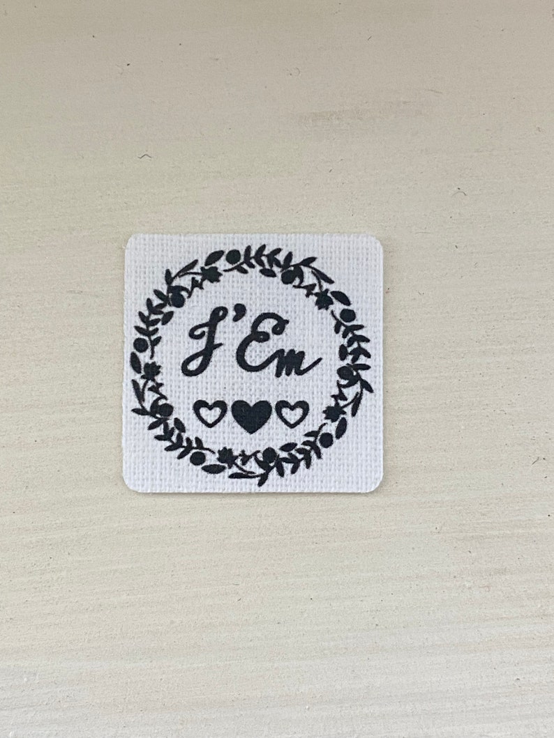 Puede incluir: Un parche cuadrado de tela blanca con texto negro y una corona decorativa. El texto dice "J'Em" dentro de una corona de hojas, con tres corazones debajo. El parche tiene una superficie texturizada.