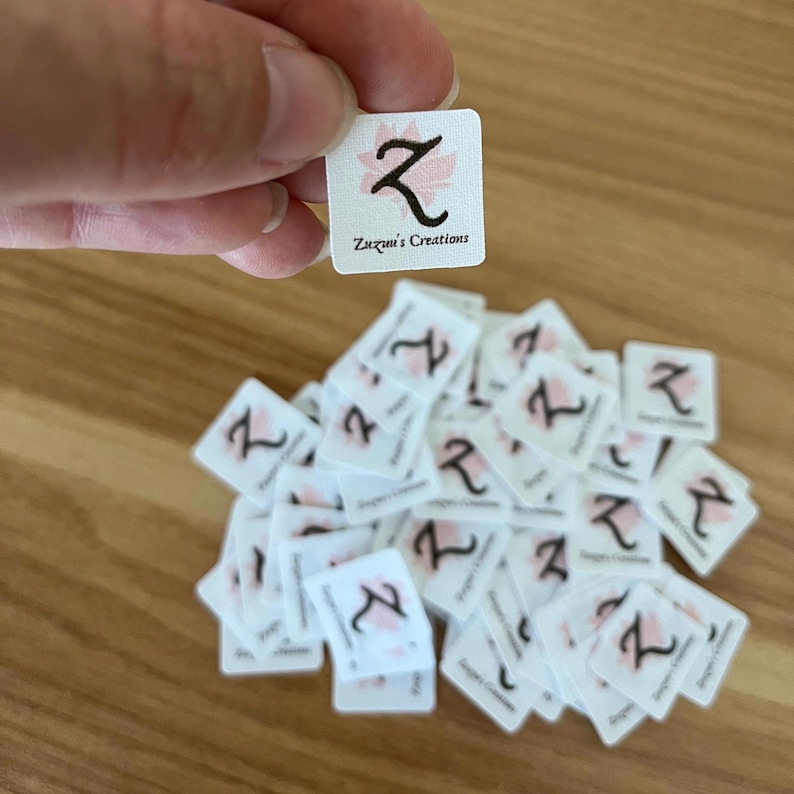 Puede incluir: Peque&ntilde;as etiquetas cuadradas blancas con un dise&ntilde;o floral rosa y una letra "Z" negra est&aacute;n esparcidas sobre una superficie de madera. Una etiqueta es sostenida por una mano. El texto "Zuzuni's Creations" est&aacute; impreso en la etiqueta.