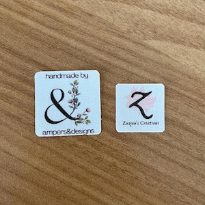 Puede incluir: Dos etiquetas cuadradas sobre una superficie de madera. La etiqueta m&aacute;s grande dice "handmade by" encima de una "&" negra con detalles florales y "ampers&designs" debajo. La etiqueta m&aacute;s peque&ntilde;a tiene una "Z" rosa y dice "Zuzuu's Creations".