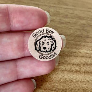Puede incluir: Una pegatina redonda de color rosa claro con las palabras "Good Boy Goodies" en negro, dispuestas en c&iacute;rculo. En el centro hay un dibujo lineal negro de la cara de un perro. La pegatina est&aacute; sujeta por los dedos de una persona.