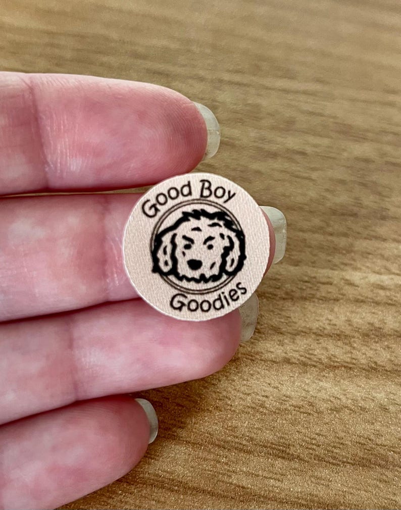 Puede incluir: Una pegatina redonda de color rosa claro con las palabras "Good Boy Goodies" en negro, dispuestas en c&iacute;rculo. En el centro hay un dibujo lineal negro de la cara de un perro. La pegatina est&aacute; sujeta por los dedos de una persona.