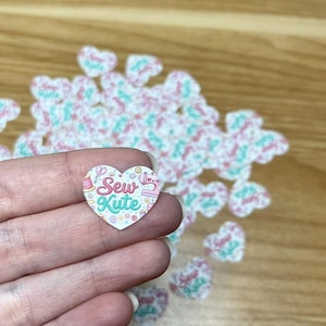 Puede incluir: Una pegatina en forma de coraz&oacute;n con el texto "Sew Kute" en una fuente caprichosa, con ilustraciones tem&aacute;ticas de costura. La pegatina se sostiene en una mano, con otras pegatinas similares dispersas en el fondo. La combinaci&oacute;n de colores incluye rosa, turquesa y blanco.