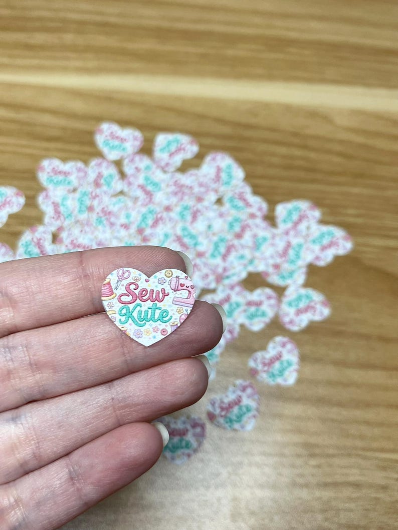 Puede incluir: Una pegatina en forma de coraz&oacute;n con el texto "Sew Kute" en una fuente caprichosa, con ilustraciones tem&aacute;ticas de costura. La pegatina se sostiene en una mano, con otras pegatinas similares dispersas en el fondo. La combinaci&oacute;n de colores incluye rosa, turquesa y blanco.