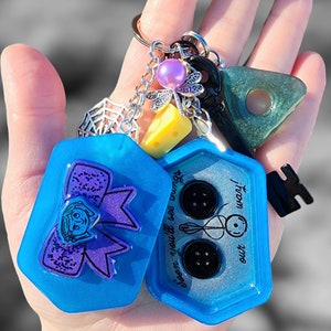 May include: A blue resin keychain with two charms. One charm features a purple bow with a girl's face and the text "Soon you'll see what we're up to". The other charm has three black buttons and the text "from our" and "little world".