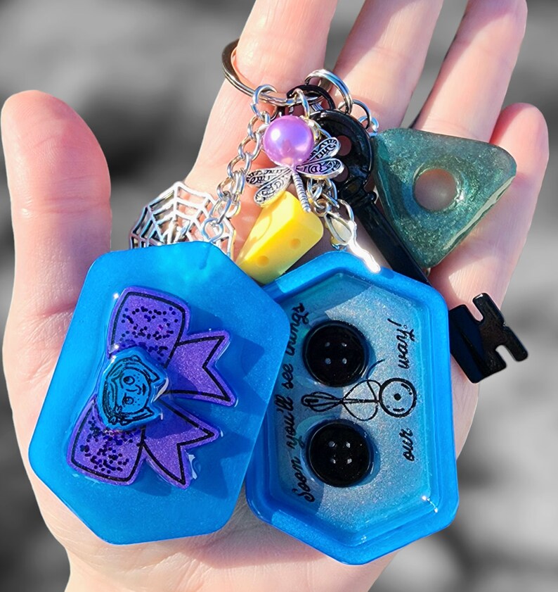 May include: A blue resin keychain with two charms. One charm features a purple bow with a girl's face and the text "Soon you'll see what we're up to". The other charm has three black buttons and the text "from our" and "little world".