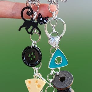 May include: Two silver-toned metal keychains featuring various charms including a black cat, a spider, a dragonfly, a turquoise triangle, a black spool of thread, a wedge of cheese, a red ball with a yellow star, and a black skeleton key.