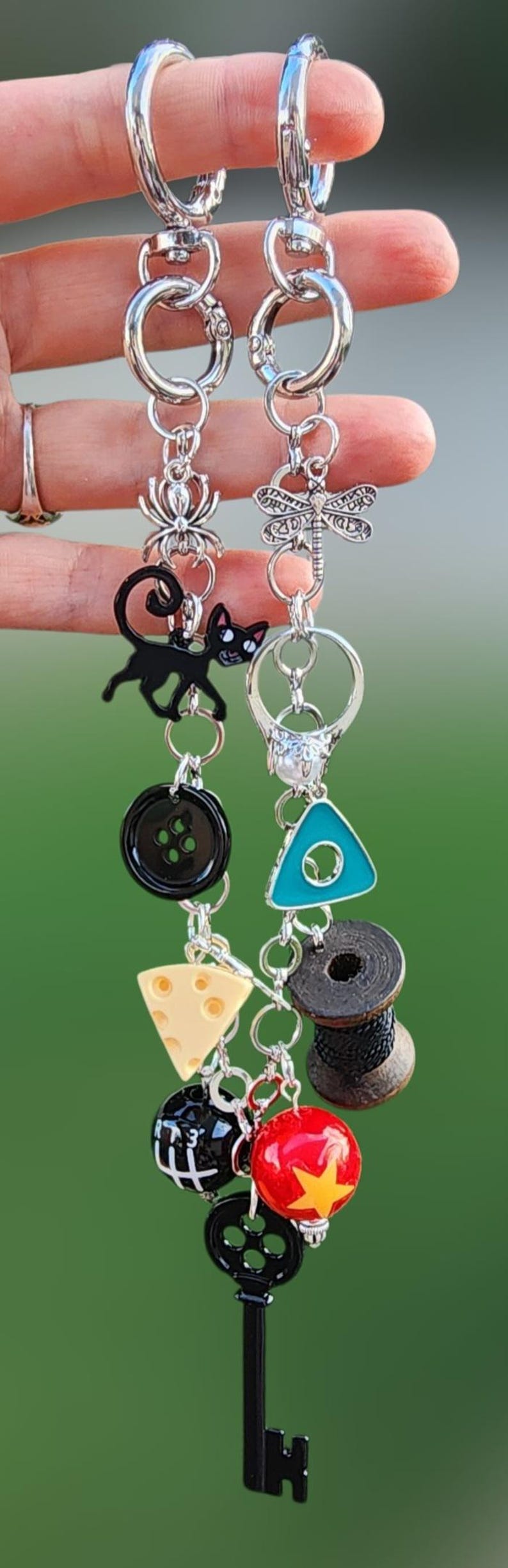 May include: Two silver-toned metal keychains featuring various charms including a black cat, a spider, a dragonfly, a turquoise triangle, a black spool of thread, a wedge of cheese, a red ball with a yellow star, and a black skeleton key.