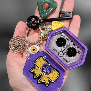 May include: Purple and gold resin keychain with a charm that says "Soon you'll see things our way!" and a charm with a bow and a girl's face. The keychain also has a spiderweb charm, a key charm, a green triangle charm, and a yellow cheese charm.
