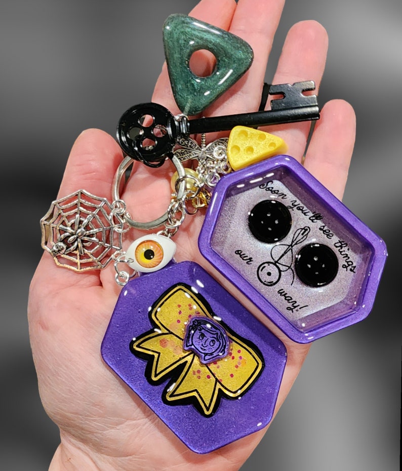 May include: Purple and gold resin keychain with a charm that says "Soon you'll see things our way!" and a charm with a bow and a girl's face. The keychain also has a spiderweb charm, a key charm, a green triangle charm, and a yellow cheese charm.