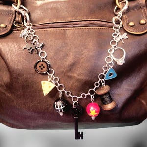 May include: A silver-toned metal charm bracelet featuring various dangling charms including a black cat, a brown button, a yellow cheese wedge, a gear shift knob, a black key, a red star bead, a thread spool, a blue triangle, and a ring.