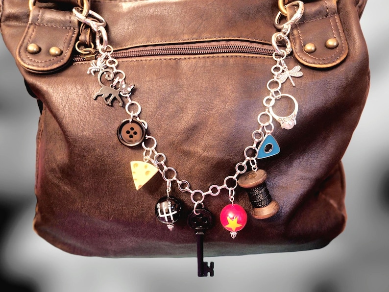 May include: A silver-toned metal charm bracelet featuring various dangling charms including a black cat, a brown button, a yellow cheese wedge, a gear shift knob, a black key, a red star bead, a thread spool, a blue triangle, and a ring.