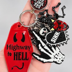 May include: A metal keychain featuring a red coffin-shaped tag with the text Highway to Hell, a circular charm with the text SEE YOU IN HELL, a black heart charm with the text SINNER, a red devil face, and a white skeleton hand charm.