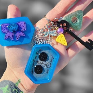 May include: A blue and silver charm keychain with a blue and purple bow, a spider web, a dragonfly, a key, a cheese wedge, and a charm with the text "Seen you'll see things with our way!"