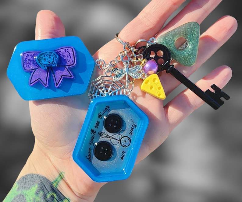 May include: A blue and silver charm keychain with a blue and purple bow, a spider web, a dragonfly, a key, a cheese wedge, and a charm with the text "Seen you'll see things with our way!"