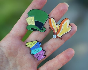 May include: Three enamel pins featuring characters from Alice in Wonderland. The first pin is a green hat with a 10/6 tag. The second pin is a stack of four teacups with the words "We Are All Mad Here". The third pin is a yellow butterfly with orange wings.