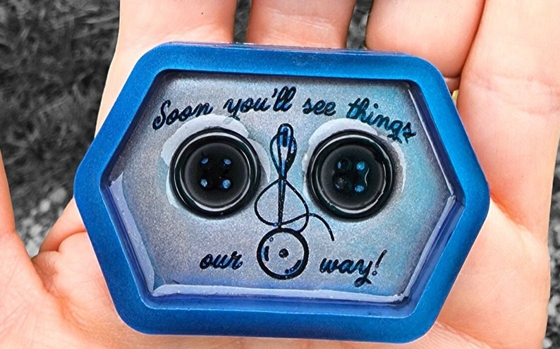 May include: A hexagonal resin tray with a blue border and a glittery silver interior. The tray features two black buttons with blue centers and a needle and thread design with the text "Soon you'll see things our way!"