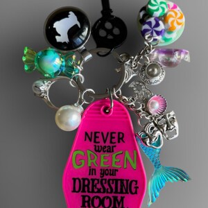 May include: A pink motel key chain with the text "NEVER wear GREEN in your DRESSING ROOM."  The keychain has a mermaid tail charm, a pearl, a candy charm, and a key charm.