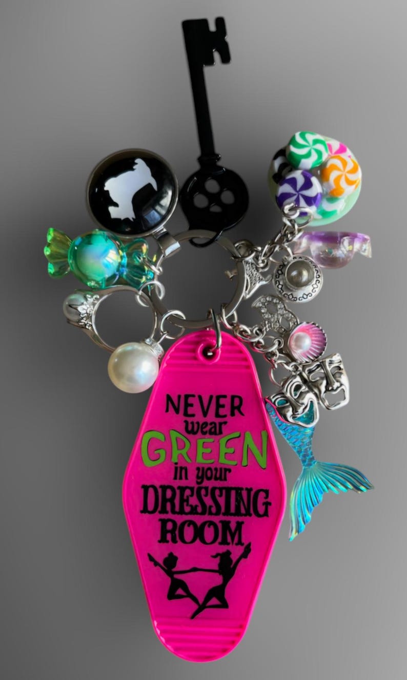 May include: A pink motel key chain with the text "NEVER wear GREEN in your DRESSING ROOM."  The keychain has a mermaid tail charm, a pearl, a candy charm, and a key charm.