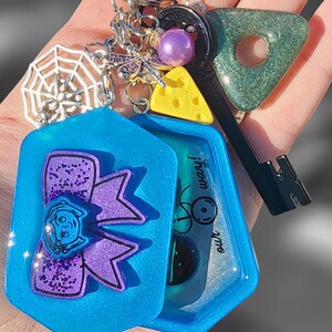 May include: A blue resin keychain with a purple glitter bow and a clear window with a green and black design. The keychain also includes a silver spiderweb charm, a silver dragonfly charm, a purple bead, a yellow cheese charm, and a black and green key charm with a triangle-shaped green charm.