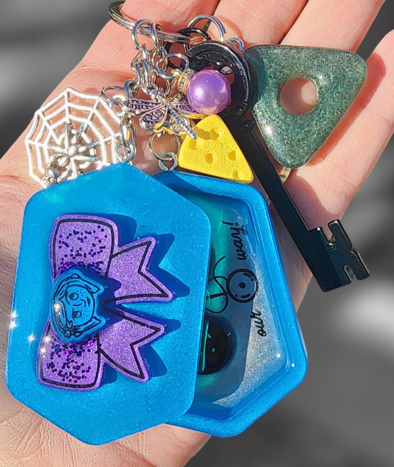 May include: A blue resin keychain with a purple glitter bow and a clear window with a green and black design. The keychain also includes a silver spiderweb charm, a silver dragonfly charm, a purple bead, a yellow cheese charm, and a black and green key charm with a triangle-shaped green charm.
