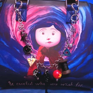 May include: A handbag featuring a Coraline character graphic with a swirling blue and pink background and the text Be careful what you wish for. A silver-toned chain is attached to the handles, adorned with charms including a black button, a slice of cheese, a black spool of thread, a red ball with a yellow star, and a black key.