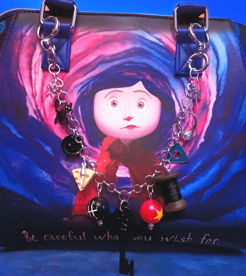 May include: A handbag featuring a Coraline character graphic with a swirling blue and pink background and the text Be careful what you wish for. A silver-toned chain is attached to the handles, adorned with charms including a black button, a slice of cheese, a black spool of thread, a red ball with a yellow star, and a black key.