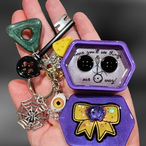 May include: A purple hexagonal resin keychain with a yellow bow and a quote that reads "Soon you'll see things our way!" The keychain also features a spider web charm with a spider, a key charm, a yellow cheese charm, and a black and white eye charm.
