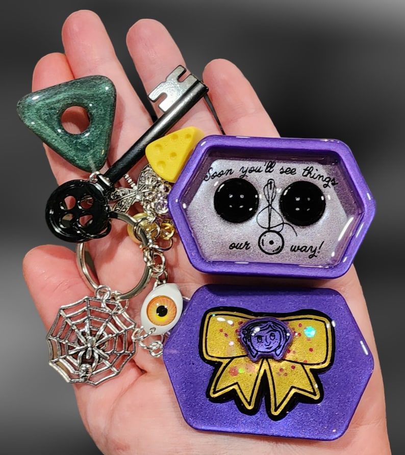 May include: A purple hexagonal resin keychain with a yellow bow and a quote that reads "Soon you'll see things our way!" The keychain also features a spider web charm with a spider, a key charm, a yellow cheese charm, and a black and white eye charm.