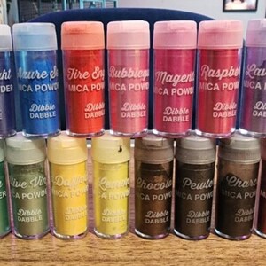 May include: A collection of 20 small jars of mica powder in various colors, including gold, blue, green, red, orange, and purple. Each jar has a white label with the name of the color and the words "Mica Powder" and "Dibble Dable".
