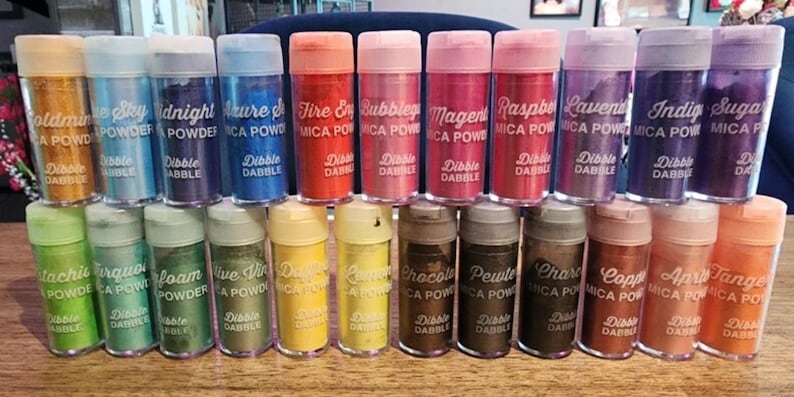 May include: A collection of 20 small jars of mica powder in various colors, including gold, blue, green, red, orange, and purple. Each jar has a white label with the name of the color and the words "Mica Powder" and "Dibble Dable".