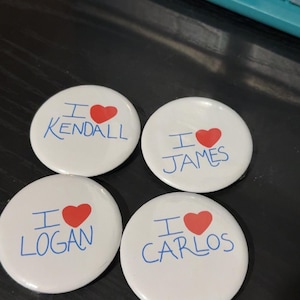 May include: Four white round buttons with blue text that says "I love" followed by a name. Each button has a red heart.
