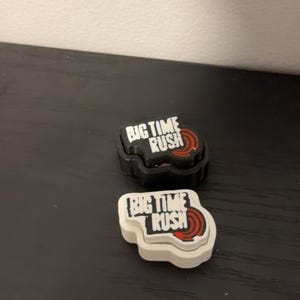 Könnte beinhalten: Zwei Schuhanhänger mit dem Schriftzug BIG TIME RUSH in weißen Buchstaben auf schwarzem Grund und einer roten kreisförmigen Grafik. Ein Anhänger hat eine schwarze Basis, der andere eine weiße. Beide haben einen welligen, unregelmäßigen Rand.