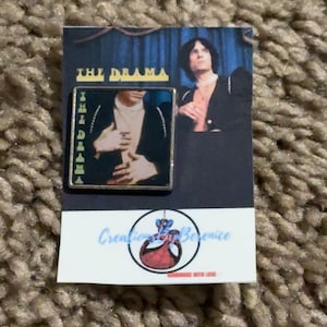 May include: A rectangular enamel pin with a portrait of a person in a black suit, with the text "THE DRAMA" at the top. The pin is on a white card with the text "Creations By Berenice".