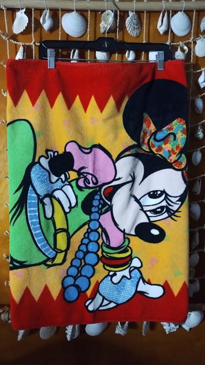 May include: A red, yellow, and green fleece blanket with a cartoon image of Minnie Mouse wearing a colorful headband and a blue and white necklace. Minnie Mouse is wearing a pink dress and has a green hat on her head.