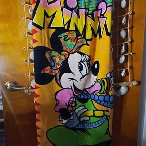 May include: A red beach towel with a yellow, green, and blue cartoon image of Minnie Mouse. The towel has the word "Minnie" written in yellow letters on it.