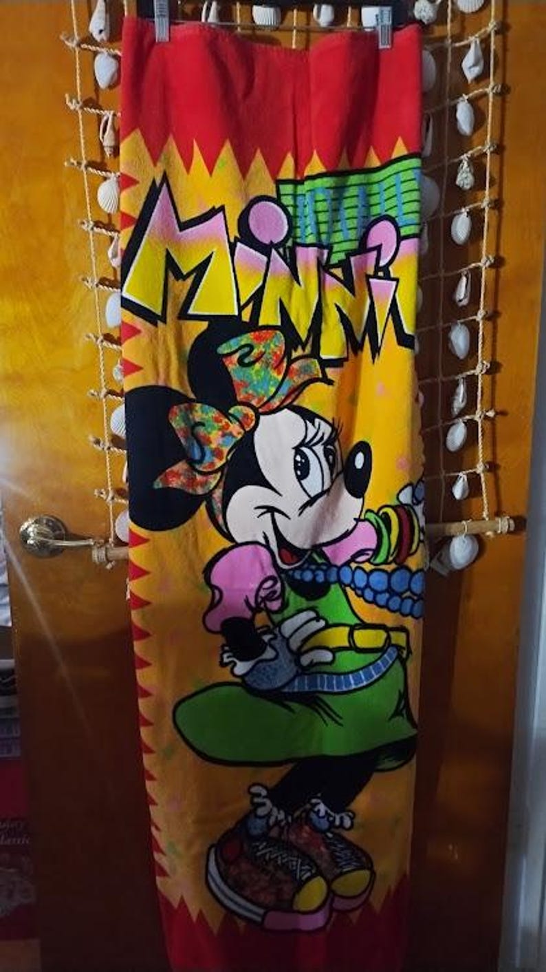 May include: A red beach towel with a yellow, green, and blue cartoon image of Minnie Mouse. The towel has the word "Minnie" written in yellow letters on it.