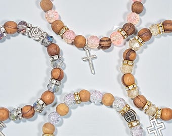 May include: Close-up of several beaded bracelets. The bracelets feature a mix of wooden, clear, and pink beads, accented with silver-toned spacers and cross charms. The beads are strung together to form a circular shape.