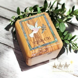 May include: Wooden box with a dove and olive branch inlay. The word "PEACE" is inscribed below the dove. The box is made of light brown wood and has a rectangular shape. The dove is white and pink, and the olive branch is green.