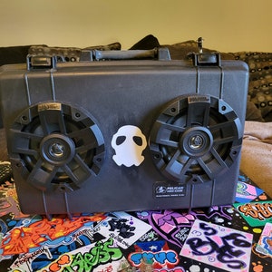 May include: A black Pelican hard case with two large speakers and a white skull sticker. The case is sitting on a table covered in colorful stickers.