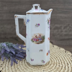 May include: A white porcelain coffee pot with a hexagonal body and a matching lid. The surface features delicate floral patterns and a central vignette depicting two figures in a landscape. The handle and rim are accented with gold trim.